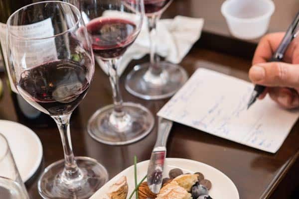 Several wine glasses filled with red wine are on a table, along with a plate of appetizers including cheese and grapes. A hand is seen writing notes on a piece of paper, likely part of a wine tasting event.