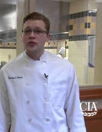 A person in a white chef's coat stands in a commercial kitchen setting. The coat has a name embroidered on it, and a small microphone is clipped to the front. The background shows kitchen equipment and another figure partially visible through a window.