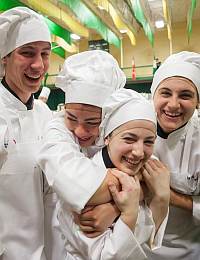 Four young chefs dressed in white uniforms and hats are laughing and embracing each other in a brightly lit kitchen. The atmosphere is joyful and energetic, with green and yellow decorations seen in the background.