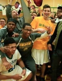 A group of young men, some wearing sports jerseys and one wearing a "Culinary" sweatshirt, gather closely and smile for the camera. They are surrounded by others who are cheering and holding up signs, creating a lively and energetic atmosphere.