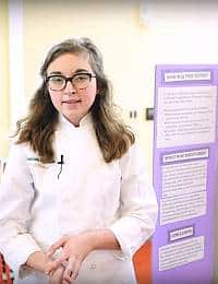 A young girl with long hair and glasses, wearing a white chef's coat, stands in front of a purple display board. The board has several sections of typed text, but they are not legible. She appears to be presenting her project.