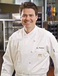 A smiling chef wearing a white chef's coat stands in a commercial kitchen. The coat has embroidered text and a small logo on the chest. The background includes industrial kitchen equipment and culinary tools.