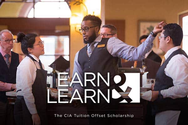 A group of culinary students in a formal dining setting, engaged in a discussion. Text overlay reads "EARN & LEARN - The CIA Tuition Offset Scholarship." A student, dressed in a vest and tie, gestures while talking to others in aprons.
