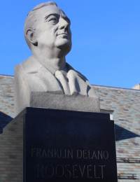 A stone bust of Franklin Delano Roosevelt, a former U.S. President, is mounted on a dark pedestal with his name engraved on the front. The background shows a building with a tiled roof and a clear blue sky.