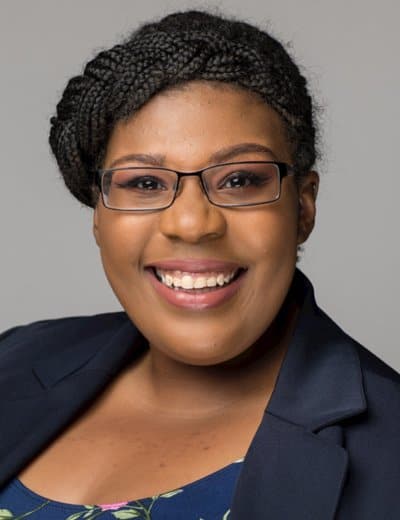 A person with braided hair, glasses, and a navy blue blazer smiles while looking at the camera. The background is plain gray.