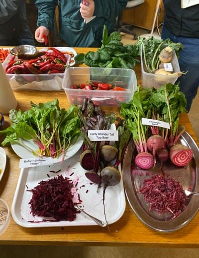 A wooden table displays various vegetables, including leafy greens, beets, and shredded red cabbage. Containers hold additional produce like red peppers. Handwritten labels identify the items, while some people in the background are partially visible but not identifiable.