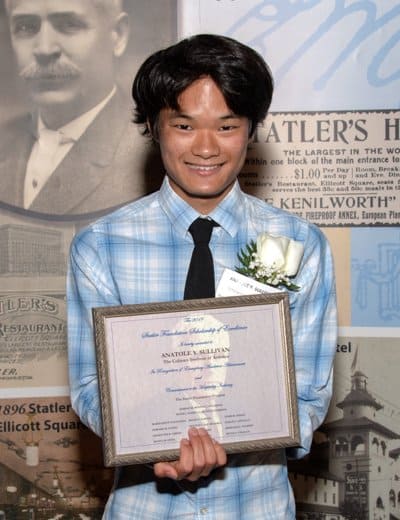 A young person with black hair smiles while holding a framed certificate. They are wearing a blue checkered shirt, black tie, and a white rose boutonniere. The background features historical images and text.