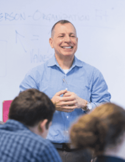 A man in a blue shirt is smiling and standing in front of a whiteboard, addressing a group of people. The whiteboard contains some writing and diagrams. The focus is on the man, who appears to be giving a presentation or a lecture.