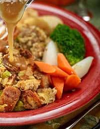 A close-up of a colorful plate filled with various foods. The plate contains stuffing, gravy being poured over the stuffing, slices of carrots, broccoli, and potato pieces. The background includes a part of a glass and a hint of a green tablecloth.