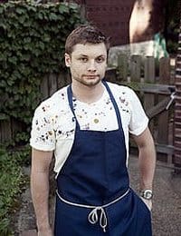 A man with short hair and a beard is standing outside wearing a white t-shirt with a splatter pattern and a dark blue apron with a white string tied at the waist. There is a wooden fence and greenery in the background.