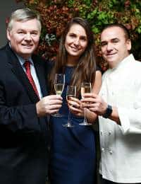 Three people are standing close together, smiling, and holding champagne glasses. On the left, a man in a suit and red tie; in the middle, a woman in a dark dress; and on the right, a man in a white chef's coat. Greenery is visible in the background.