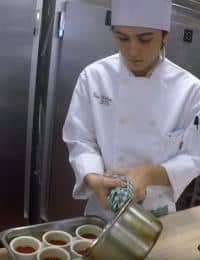 A chef in a white uniform and hat is carefully pouring liquid from a pot into small ceramic containers. He is standing in a kitchen with stainless steel appliances and holding a cloth in his left hand.