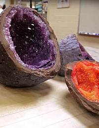 An image showing two geodes cut open to reveal their crystal interiors. The larger geode has purple amethyst crystals, while the smaller one displays vibrant orange crystals. Both geodes are resting on a wooden surface.