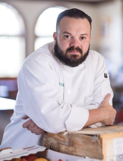 A man with a beard wearing a white chef's coat leans on a wooden surface. He is looking directly into the camera with a serious expression. Blurred background features arched windows allowing light to stream in, creating a soft and warm atmosphere.