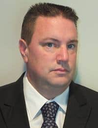 A person with short brown hair, wearing a white dress shirt, dark tie, and dark suit jacket, looks slightly to the right with a serious expression in a professional portrait.