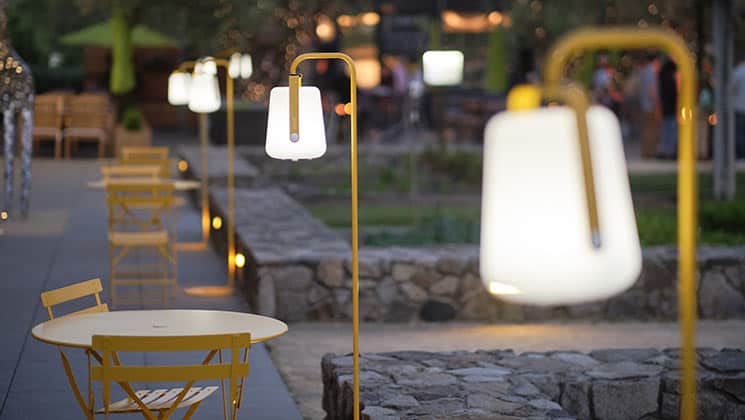 A serene outdoor café setting with yellow chairs and tables on a stone-paved area. Brightly lit lamps mounted on yellow stands line the area, casting a warm glow. Stone walls and blurred greenery appear in the background, adding to the tranquil ambiance.