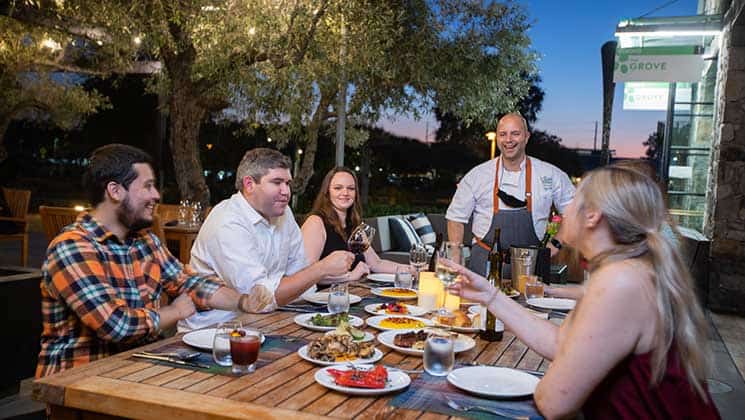 A group of friends is seated around a wooden table enjoying a meal in an outdoor dining area during the evening. A chef stands nearby, smiling. The table is covered with various dishes and drinks, and the setting is illuminated with soft, warm lighting.