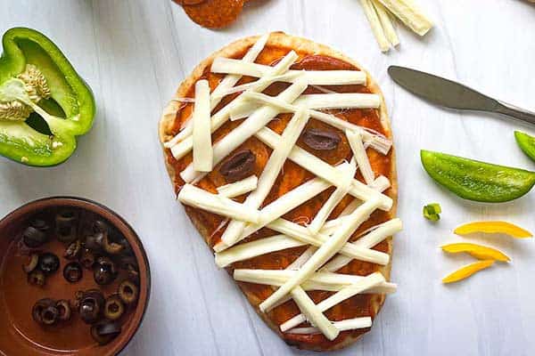 A mummy-themed pizza on a white surface is topped with strips of cheese resembling bandages and olive slices for eyes. Surrounding the pizza are ingredients including a green bell pepper, a small bowl of sliced olives, a knife, and some yellow pepper slices.