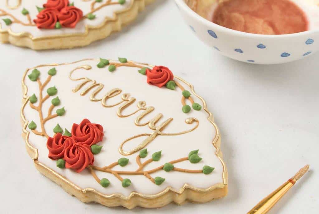 A decorated cookie shaped like an ornate plaque with the word "merry" written in gold icing. The cookie is adorned with red rose-like flowers and green leaves. A small paintbrush and a bowl containing gold luster dust are nearby.
