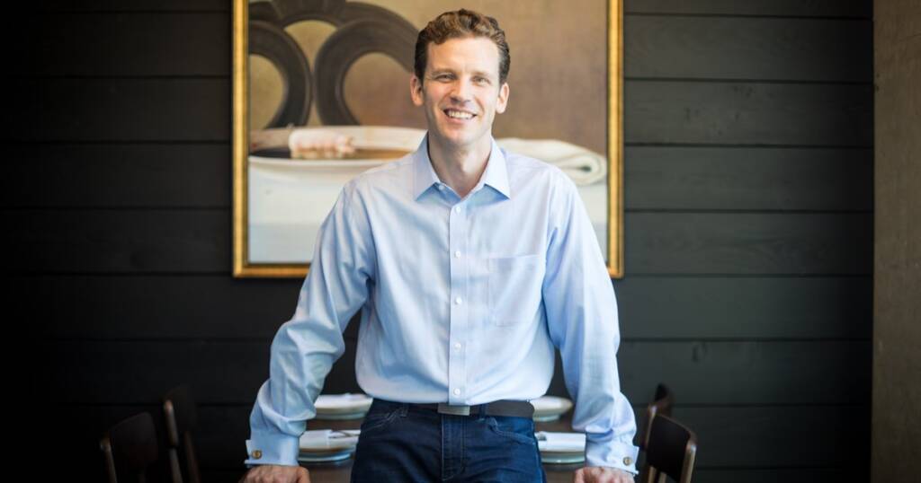 A man wearing a light blue dress shirt and dark blue jeans leans against a table, smiling. The background features a dark wooden wall with a framed painting of plates and napkins. Two chairs are partially visible on either side of him.