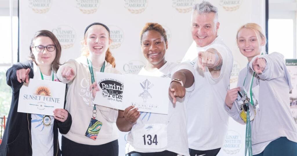 A group of five people, all smiling, stand pointing towards the camera. They are wearing event medals and <a href=