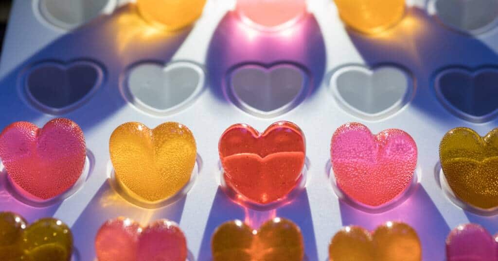 A close-up image of heart-shaped translucent gummies, with colors of yellow, pink, and red, arranged neatly in a mold or tray. The light casts shadows and highlights the texture of the gummies, emphasizing their glossy and chewy appearance.