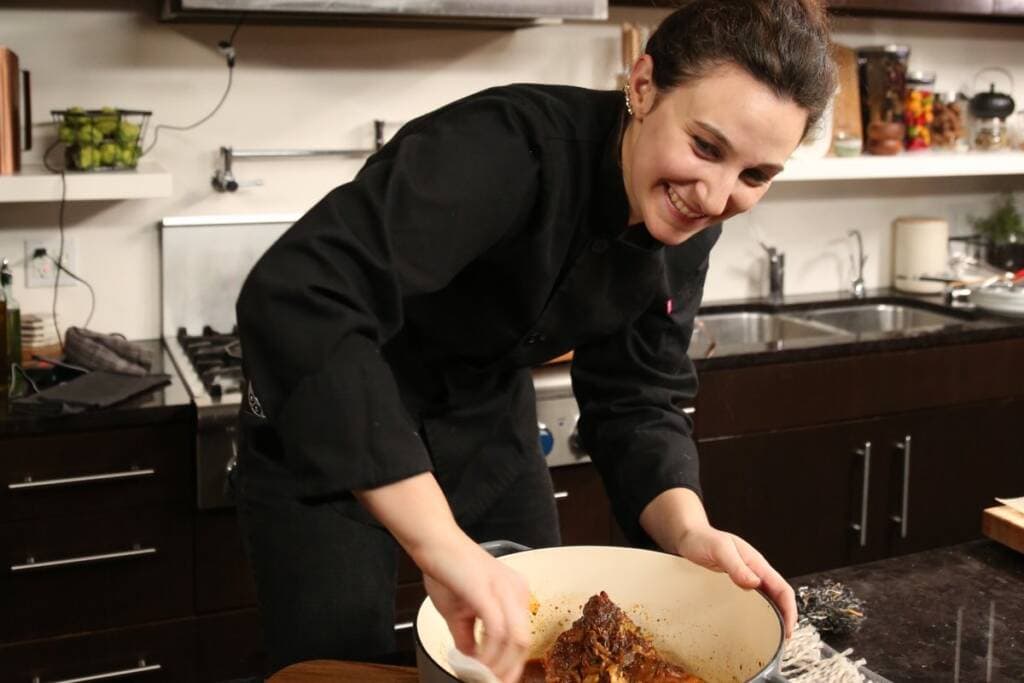 A chef in a black uniform stirs a pot of food in a modern kitchen. The chef is smiling and looking at the dish while cooking. The kitchen has various utensils, a gas stove, and ingredients visible in the background.