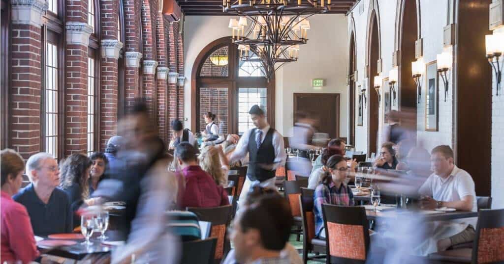 A bustling restaurant with numerous patrons sitting at tables, dining and conversing. Waitstaff, captured in motion, are serving food and drinks. The environment features brick walls, arched windows, and elegant chandeliers, creating a warm and inviting atmosphere.