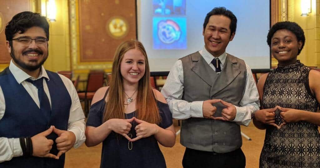 Four people stand in a row, smiling. Two men and two women are making similar hand gestures, forming a triangle with their fingers. The background shows a room with a projector screen and chairs. The people are wearing semi-formal attire.