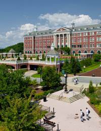 A large, elegant red-brick building with white columns stands against a backdrop of a blue sky with scattered clouds. A well-manicured lawn, trees, a fountain, walking paths, and a few people are visible in the foreground.