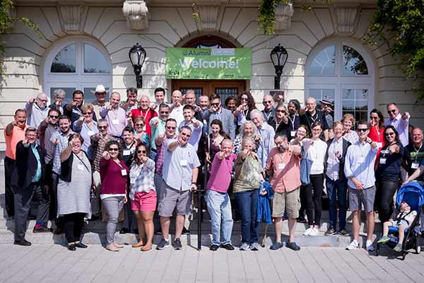 A large group of people, likely alumni, pose for a group photo in front of a building with a green banner that reads, "Welcome!" Many smile and wave at the camera, standing on steps and beneath a few outdoor lamps flanking the entrance.