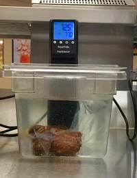 A sous vide machine cooking a piece of meat submerged in water in a clear container. The machine's digital display shows a temperature of 125°F and a timer reading 1:70. The setup is placed on a countertop.