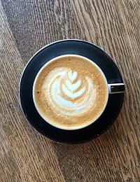 A cup of cappuccino with a heart-shaped latte art design on the foam sits on a black saucer on a wooden table.