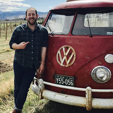 A man stands smiling and pointing to a vintage red Volkswagen van in a scenic outdoor location with mountains and a vineyard in the background. The van has an old license plate and a prominent Volkswagen logo on the front.