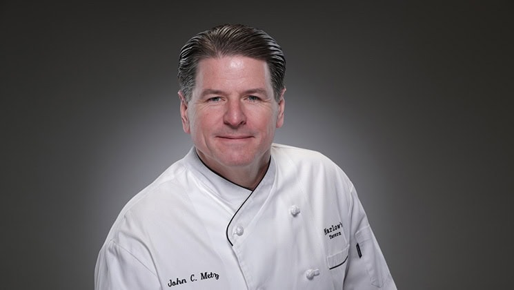 A middle-aged man with neatly combed hair, wearing a white chef's jacket, smiles at the camera. His name is embroidered on the jacket along with the word "Executive Chef". The background is a gradient from dark to light gray.