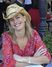 A woman with blonde hair, wearing a straw cowboy hat and a red patterned blouse, smiles while sitting at an outdoor table with her arms crossed. There are people in the background and some greenery nearby.