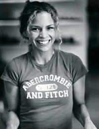 A woman with curly hair tied back smiles widely at the camera. She is wearing a T-shirt with the text "Abercrombie and Fitch" printed on it. The image is in black and white, and the background is slightly blurred.