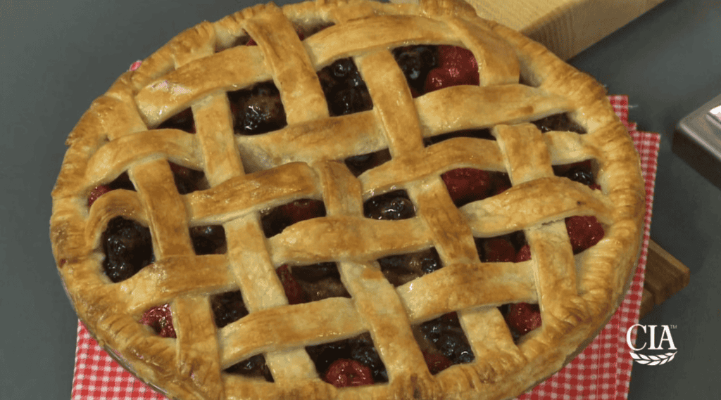 A freshly baked pie with a golden-brown lattice crust, filled with mixed dark and red berries, is placed on a red and white checkered cloth. The pie appears to be sitting on a wooden board. The bottom right corner features a small logo with the letters "CIA" and a laurel wreath.