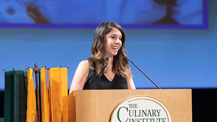 A woman wearing a black sleeveless top is standing behind a podium with the emblem of The Culinary Institute, speaking into a microphone. Colorful banners are displayed to her left. The background is a blurred projection screen.