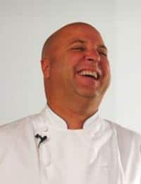 A bald man wearing a white chef's coat is laughing heartily. The background is plain and white, and a small microphone is clipped to the front of his coat.