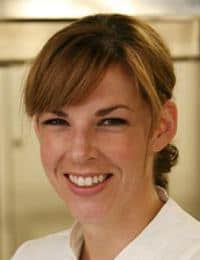 A smiling woman with light brown hair, tied back, and bangs, is wearing a white shirt. The background is blurred and appears to be an indoor setting.