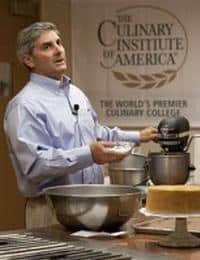 A man in a blue shirt appears to be giving a cooking demonstration at The Culinary Institute of America. He is holding a white bowl and standing behind a large metallic mixing bowl with a cake and utensils in front of him. The institute's logo is visible in the background.