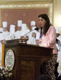 A woman wearing a pink jacket speaks at a podium adorned with a seal reading "Culinary Institute of America." In the background, chefs in white uniforms and hats are seated.