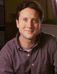 A person with short dark hair, wearing a dark button-up shirt over a lighter t-shirt, is smiling at the camera against an indoor background. The setting appears to be softly lit.