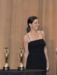 A woman in a black dress stands beside a table with two golden trophies. She is smiling and looking to her right. The background features brown drapes.
