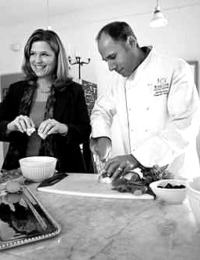 A black-and-white photo shows a woman and a man in a kitchen. The woman, smiling, is cracking an egg into a bowl. The man, dressed in a chef's coat, is chopping vegetables on a cutting board. Various kitchen items are arranged on the counter in front of them.