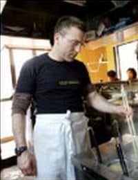 A man wearing glasses, a black shirt, and a white apron is standing in a kitchen. He is looking down at a cooking area where utensils are placed. The background shows some kitchen equipment and a window with natural light streaming in.