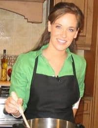 A person with long brown hair, wearing a green shirt and a black apron, smiles while stirring food in a pot in a kitchen. The background shows wooden cabinets and tiled walls.