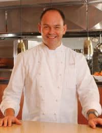 A smiling chef in a white double-breasted chef's coat stands in a kitchen with stainless steel accents. The chef is positioned in front of a counter, with their hands resting on it, and there are hanging lamps and kitchen utensils in the background.