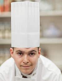 A chef wearing a tall white chef's hat and a white uniform looks directly at the camera with a slight smile. The background appears to be a blurred kitchen setting with various ingredients and items on shelves.